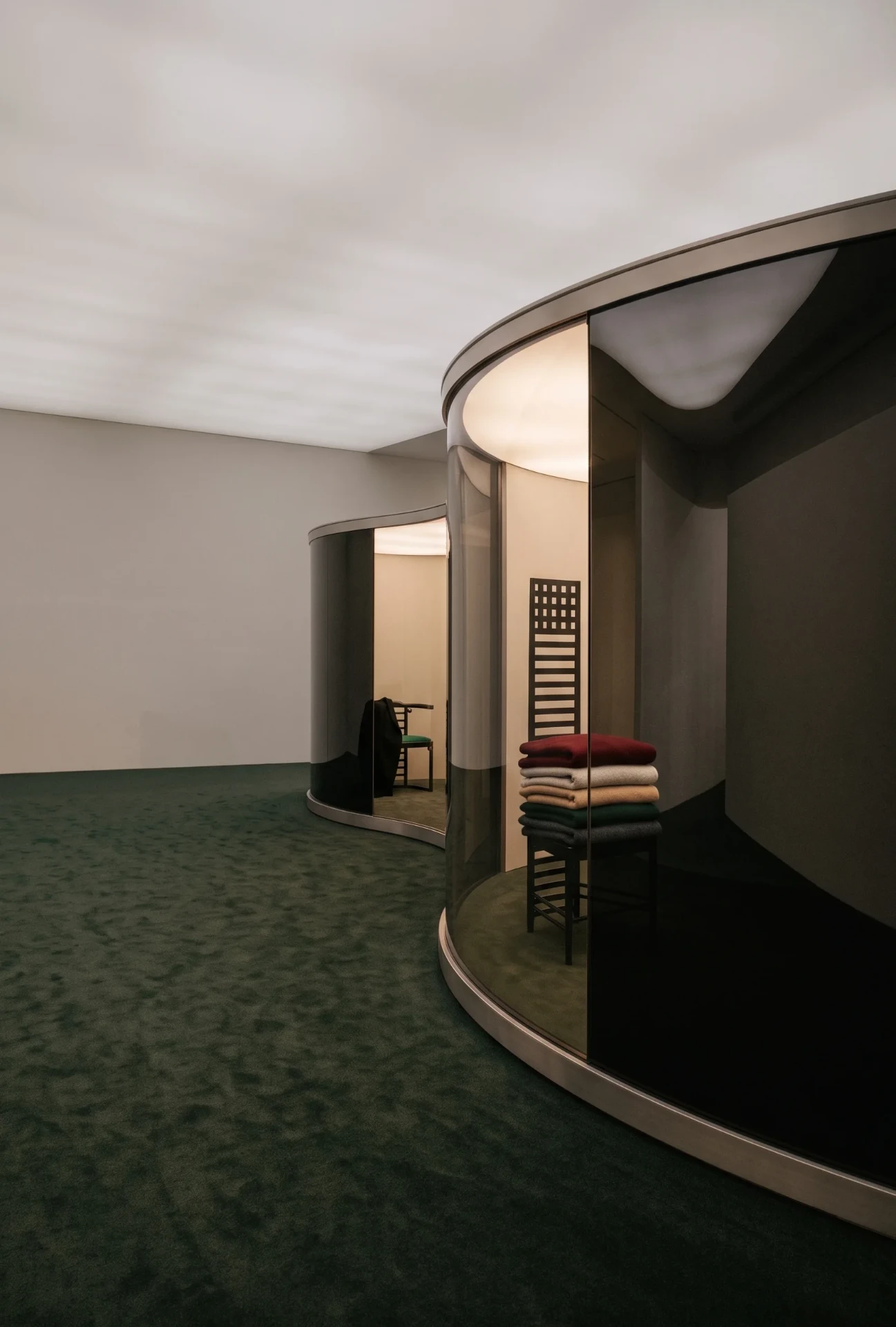 Glass pavilions reveal chairs and textiles as the room dims.