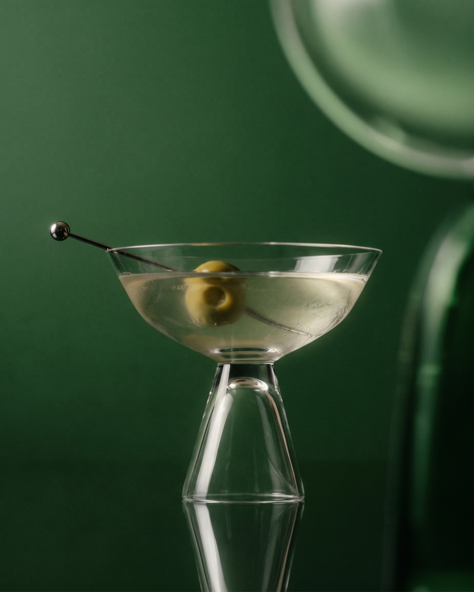 Martini glass with an olive on a cocktail pick against a deep green backdrop.