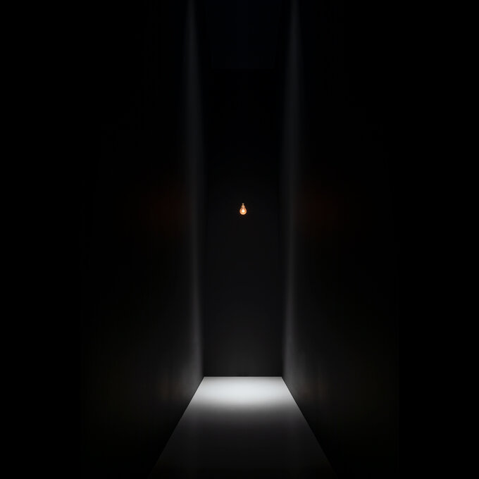 A single glowing bulb suspended in a tall dark corridor above a lit patch of floor.